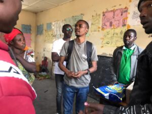 a young black man in a grey shirt and jeans speaking to a group of people (unseen)